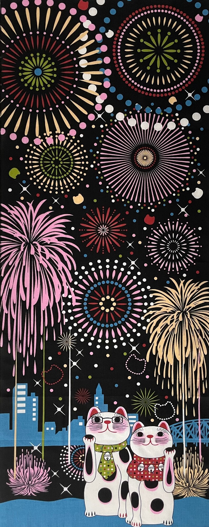 May include: Two white lucky cat figurines with black spots, wearing green and red collars with bells, stand in front of a cityscape with fireworks exploding in the night sky. The fireworks are pink, red, yellow, and blue.
