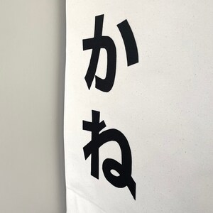 Puede incluir: Una pancarta de lona blanca con los caracteres japoneses "か ね" impresos en negro.