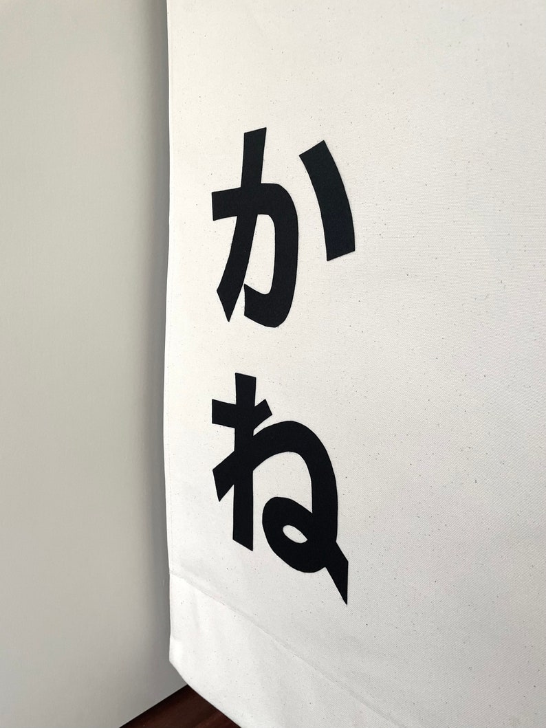 Puede incluir: Una pancarta de lona blanca con los caracteres japoneses "か ね" impresos en negro.
