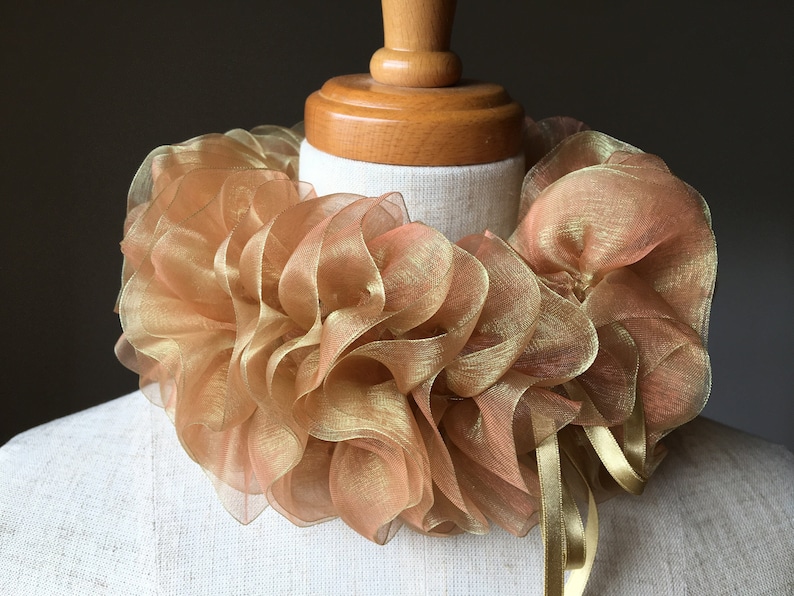 Puede incluir: Un cuello dorado con volantes de tela transparente. El cuello se muestra en un maniqu&iacute; y presenta un dise&ntilde;o en capas con lazos de cinta. La tela tiene una apariencia brillante y met&aacute;lica.