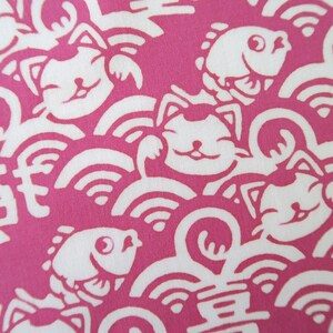 Pu&ograve; includere: Un motivo in tessuto con un design ripetitivo di gatti portafortuna e pesci bianchi su uno sfondo magenta vibrante. I gatti sono raffigurati con le zampe alzate e volti sorridenti, intervallati da motivi a onde stilizzati e caratteri kanji.