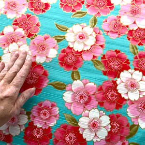 May include: A teal fabric with a repeating pattern of pink and white flowers with gold accents. The flowers have a layered petal design.