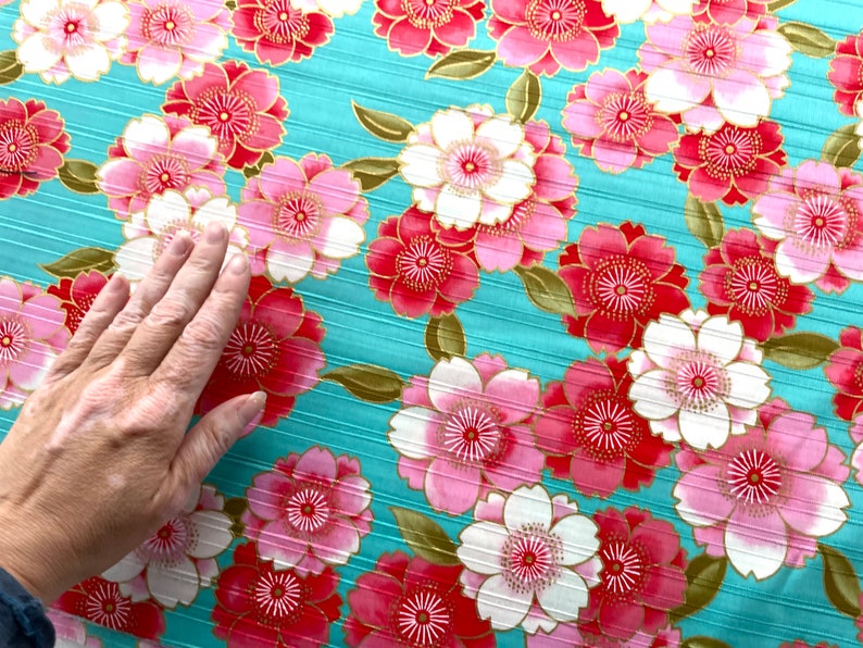 May include: A teal fabric with a repeating pattern of pink and white flowers with gold accents. The flowers have a layered petal design.