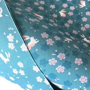 Peut inclure: Gros plan sur deux &eacute;chantillons de tissu. L'un est bleu clair uni, l'autre est vert d'eau avec un motif r&eacute;p&eacute;t&eacute; de fleurs blanches, de lapins au ventre rose et de formes florales bleues stylis&eacute;es. Le tissu est probablement du coton ou une mati&egrave;re similaire.