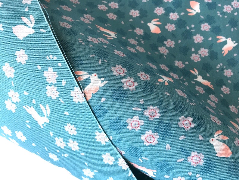 Peut inclure: Gros plan sur deux &eacute;chantillons de tissu. L'un est bleu clair uni, l'autre est vert d'eau avec un motif r&eacute;p&eacute;t&eacute; de fleurs blanches, de lapins au ventre rose et de formes florales bleues stylis&eacute;es. Le tissu est probablement du coton ou une mati&egrave;re similaire.