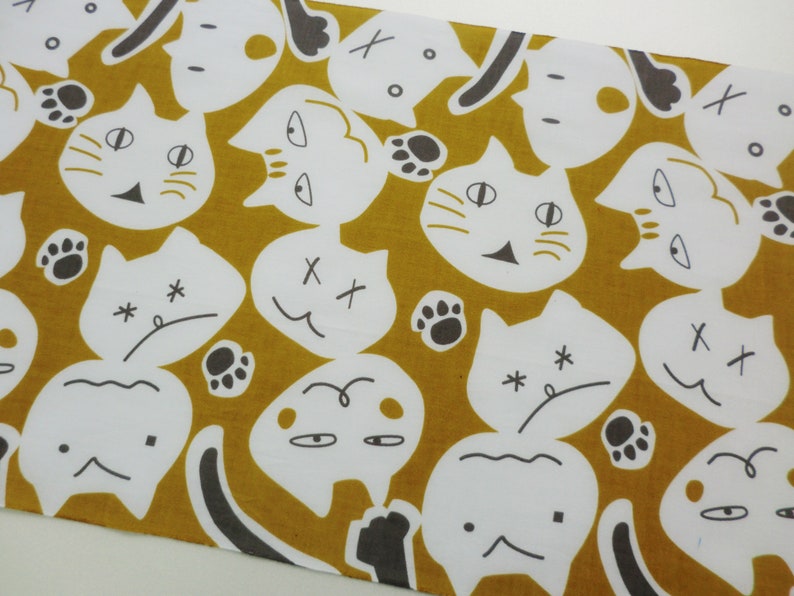 May include: A mustard yellow fabric with a repeating pattern of white cats with different facial expressions. The cats have black eyes, noses, and whiskers. Some cats have X's for eyes, while others have closed eyes. Some cats have paw prints on their heads.