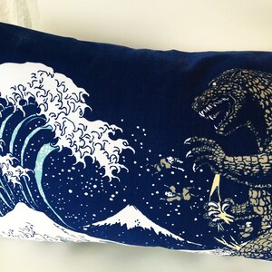 May include: A blue pillow with a white wave and a brown Godzilla-like monster printed on it. The wave is a stylized version of the Great Wave off Kanagawa, a famous Japanese woodblock print.