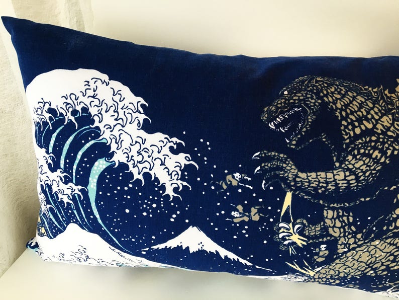 May include: A blue pillow with a white wave and a brown Godzilla-like monster printed on it. The wave is a stylized version of the Great Wave off Kanagawa, a famous Japanese woodblock print.