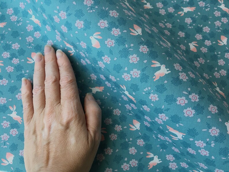Peut inclure: Tissu turquoise avec un motif r&eacute;p&eacute;t&eacute; de lapins et de fleurs roses. Le tissu a un aspect textur&eacute; avec un motif de grille subtil. Les lapins sont repr&eacute;sent&eacute;s en train de courir, ajoutant une touche fantaisiste au textile.