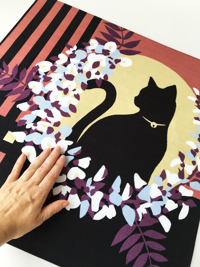 以下が含まれることがあります： 黒猫のシルエットが、金色の首輪をつけて、白と青の藤の花の輪の中に座っています。背景は黒で、右上の角にはピンクと黒のストライプ模様があります。