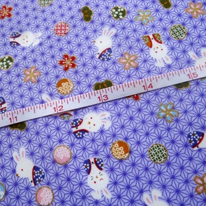 May include: Close-up of a lavender fabric with a repeating pattern of white rabbits, floral designs, and geometric shapes. A measuring tape is laid across the fabric, showing centimetre measurements. The fabric has a Japanese aesthetic.