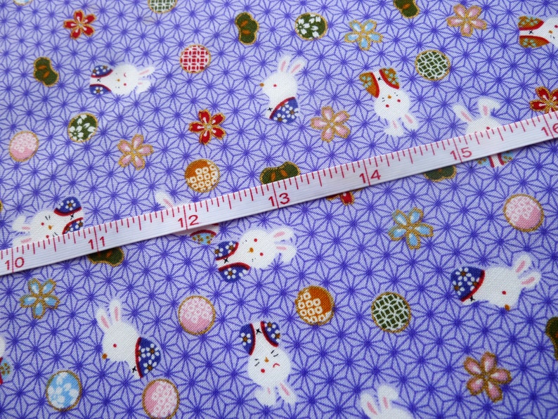 May include: Close-up of a lavender fabric with a repeating pattern of white rabbits, floral designs, and geometric shapes. A measuring tape is laid across the fabric, showing centimetre measurements. The fabric has a Japanese aesthetic.