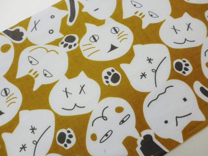 May include: A mustard yellow fabric with a white repeating pattern of cat faces. The cat faces have different expressions, including happy, sad, and surprised. Some of the cat faces have a single X on their foreheads, while others have two Xs. Some of the cat faces have paw prints next to them.