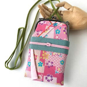 May include: A rectangular fabric pouch with a dark metal kiss-lock clasp and a long green strap. The pouch features a pink floral pattern with white grid accents, a wide teal band across the center, and a thin pink cord tied in a knot.