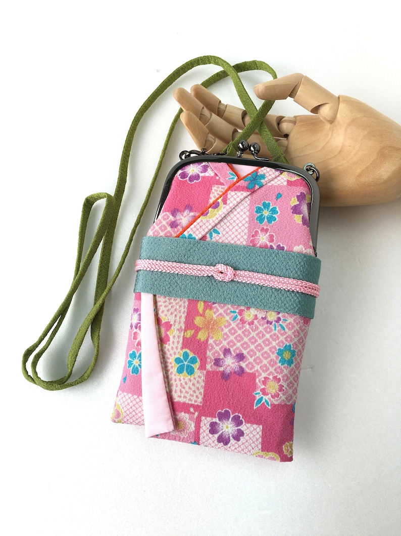 May include: A rectangular fabric pouch with a dark metal kiss-lock clasp and a long green strap. The pouch features a pink floral pattern with white grid accents, a wide teal band across the center, and a thin pink cord tied in a knot.