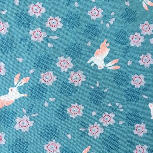 Peut inclure: Tissu turquoise avec un motif r&eacute;p&eacute;t&eacute; de lapins blancs aux accents p&ecirc;che, de fleurs blanches et rouges et de formes g&eacute;om&eacute;triques bleues. Le tissu a un aspect textur&eacute;.