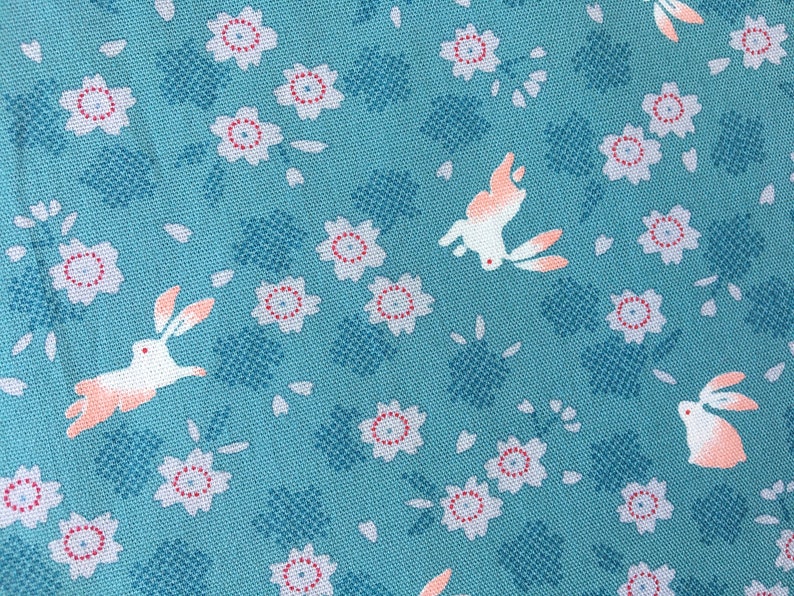 Peut inclure: Tissu turquoise avec un motif r&eacute;p&eacute;t&eacute; de lapins blancs aux accents p&ecirc;che, de fleurs blanches et rouges et de formes g&eacute;om&eacute;triques bleues. Le tissu a un aspect textur&eacute;.