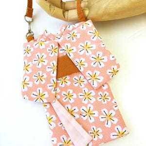 May include: A small, rectangular pouch shaped like a kimono, crafted from a light pink fabric featuring a repeating white and yellow cherry blossom pattern. It includes a solid brown fabric sash across the center and a thin brown strap with a metal clasp.