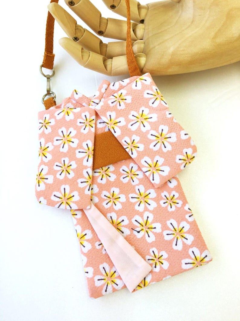 May include: A small, rectangular pouch shaped like a kimono, crafted from a light pink fabric featuring a repeating white and yellow cherry blossom pattern. It includes a solid brown fabric sash across the center and a thin brown strap with a metal clasp.