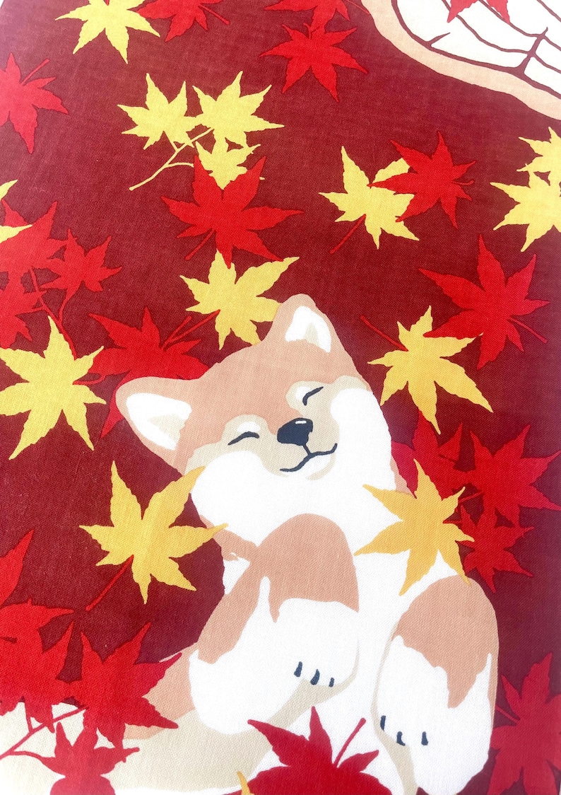 May include: A fabric textile featuring a tan and white Shiba Inu dog with a happy expression, surrounded by scattered red and yellow maple leaves against a deep maroon background. The dog is depicted lying down with its eyes closed and paws raised.