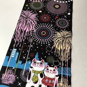 May include: Two white lucky cats with black spots and pink cheeks are standing in front of a cityscape with fireworks exploding in the night sky. The cats are wearing scarves and one has a green tie. The background is black with white stars and the cityscape is blue.