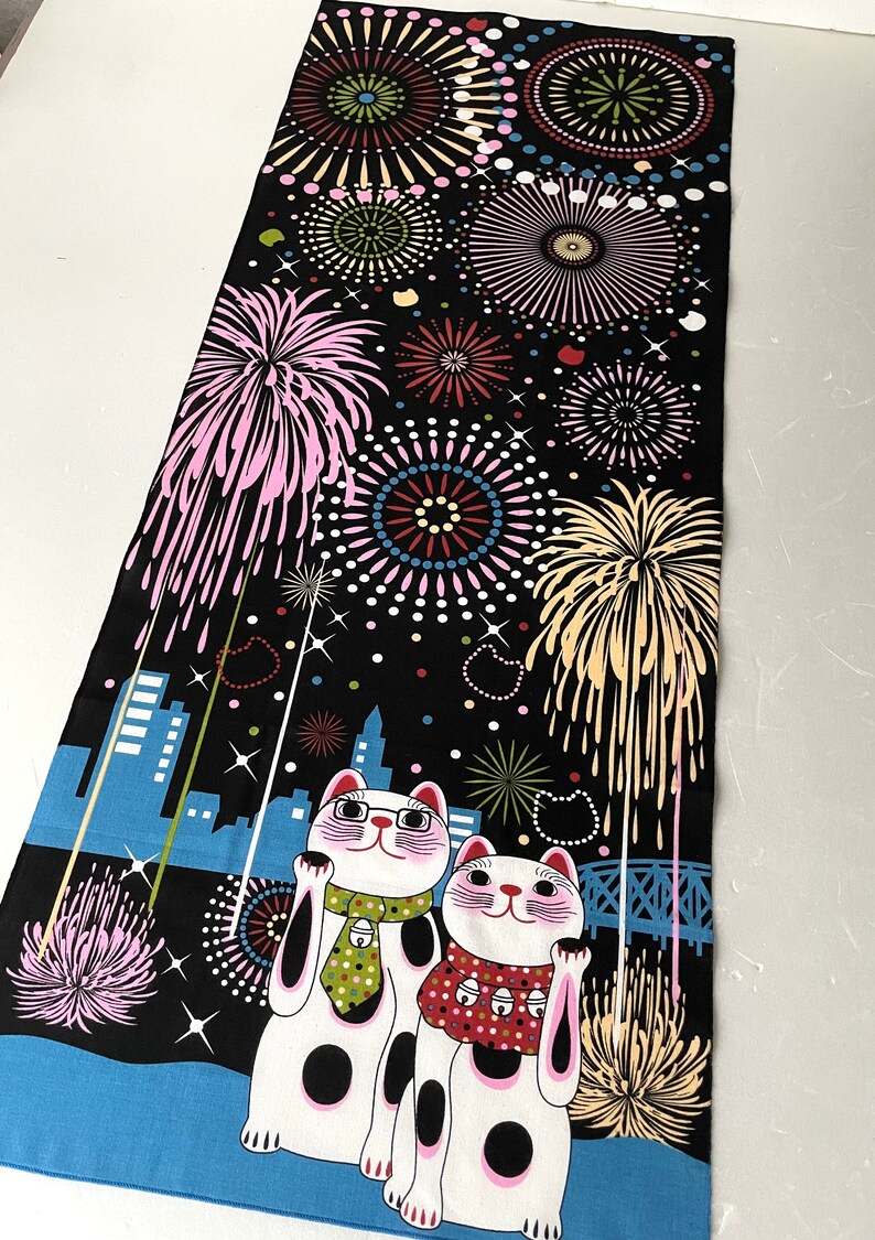 May include: Two white lucky cats with black spots and pink cheeks are standing in front of a cityscape with fireworks exploding in the night sky. The cats are wearing scarves and one has a green tie. The background is black with white stars and the cityscape is blue.