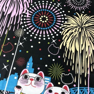 May include: Two white lucky cat figurines with black eyes and pink cheeks, wearing green and red scarves, are standing in front of a cityscape with fireworks in the background. The fireworks are in various colors, including pink, yellow, and blue.