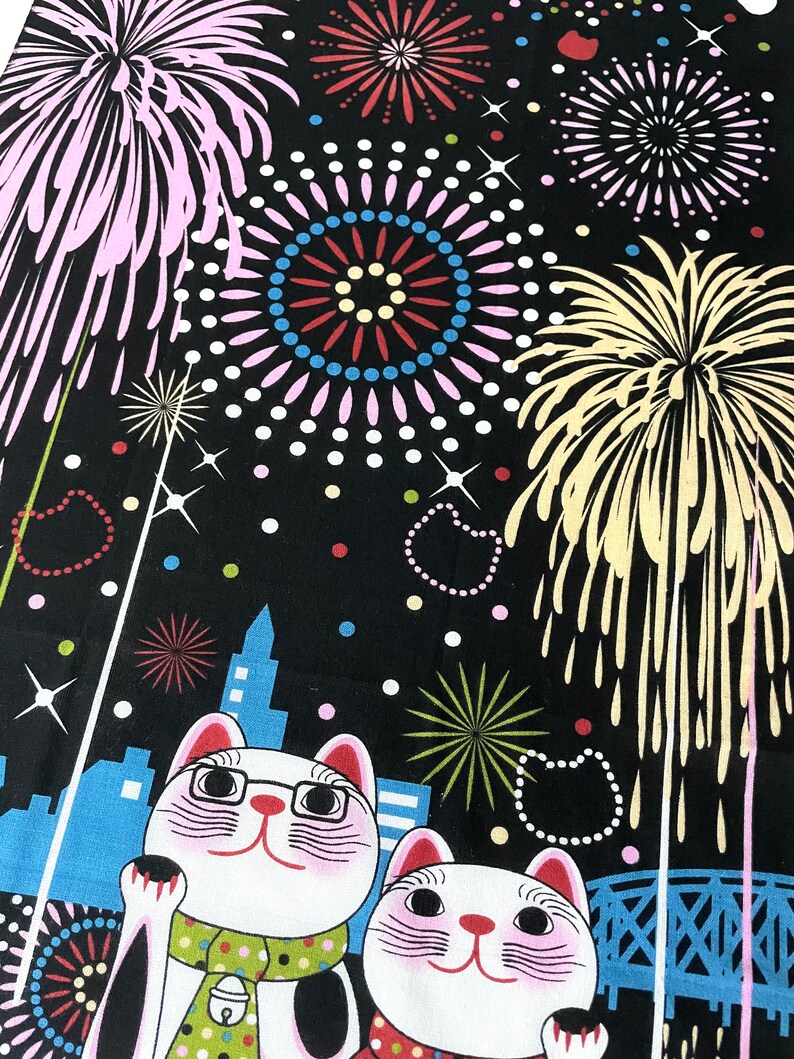 May include: Two white lucky cat figurines with black eyes and pink cheeks, wearing green and red scarves, are standing in front of a cityscape with fireworks in the background. The fireworks are in various colors, including pink, yellow, and blue.