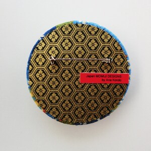 May include: A circular brooch featuring a black and gold hexagonal pattern with a floral motif. The back includes a silver metal pin mechanism and a red rectangular label with the text Japan MOMIJI DESIGNS by Ana Kondo printed in black.