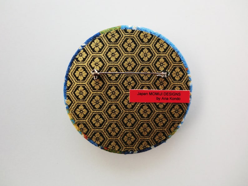 May include: A circular brooch featuring a black and gold hexagonal pattern with a floral motif. The back includes a silver metal pin mechanism and a red rectangular label with the text Japan MOMIJI DESIGNS by Ana Kondo printed in black.