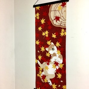 May include: A vertical fabric wall hanging featuring a Shiba Inu dog lying on its back amidst a pattern of red and yellow maple leaves on a dark red background. The textile is suspended from a black wooden frame with a black string for hanging.