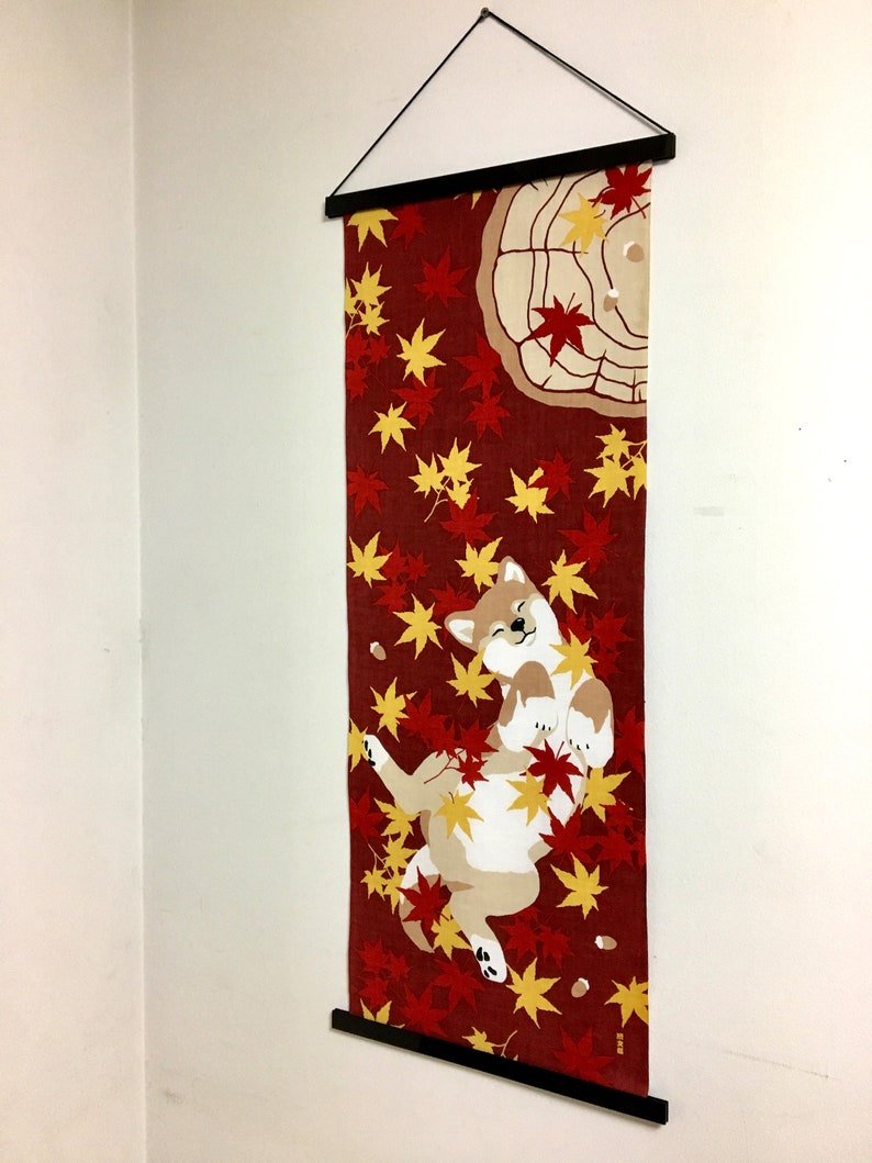 May include: A vertical fabric wall hanging featuring a Shiba Inu dog lying on its back amidst a pattern of red and yellow maple leaves on a dark red background. The textile is suspended from a black wooden frame with a black string for hanging.