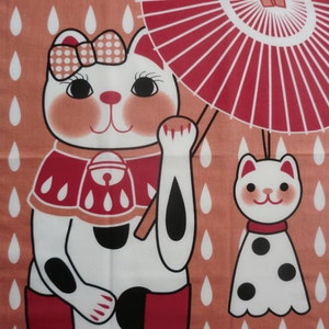 May include: A white cat with black spots and red boots holds a red and white umbrella. The cat is wearing a red and white bib with a bell. The background is a peach color with white raindrops. A smaller white cat with black spots is standing to the right of the larger cat.