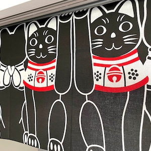 Puede incluir: Cortina de estilo japon&eacute;s en blanco y negro con un patr&oacute;n repetido de gatos negros estilizados con collares rojos y blancos. Los gatos tienen caras sonrientes y est&aacute;n en una pose de bienvenida. La cortina cuelga de una barra plateada.
