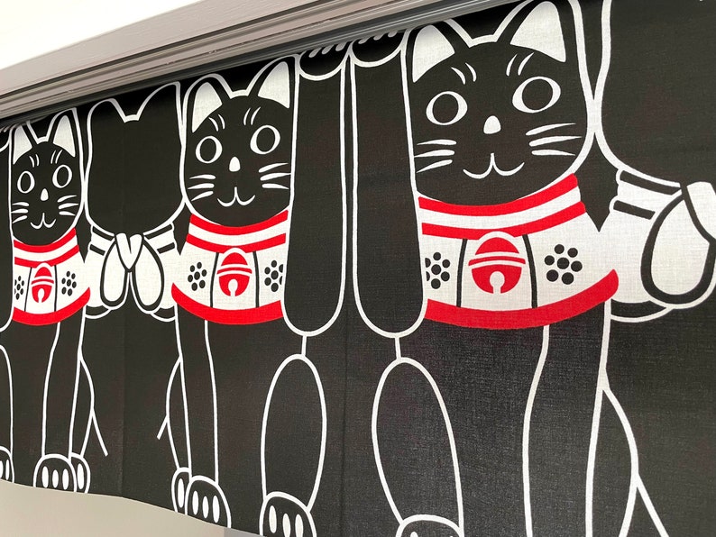 Puede incluir: Cortina de estilo japon&eacute;s en blanco y negro con un patr&oacute;n repetido de gatos negros estilizados con collares rojos y blancos. Los gatos tienen caras sonrientes y est&aacute;n en una pose de bienvenida. La cortina cuelga de una barra plateada.
