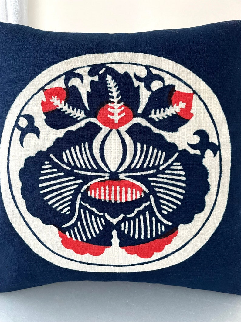 Op de afbeelding: Een donkerblauw kussen met een wit cirkelvormig ontwerp met een gestileerde rode en zwarte bloem.