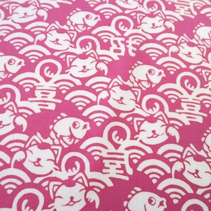 Pu&ograve; includere: Un tessuto rosa vivace con un motivo bianco ripetuto di gatti portafortuna stilizzati, pesci e onde. Il design include piccoli caratteri cinesi intervallati tra i gatti e i pesci, creando una stampa grafica densa e continua su tutto il tessuto.