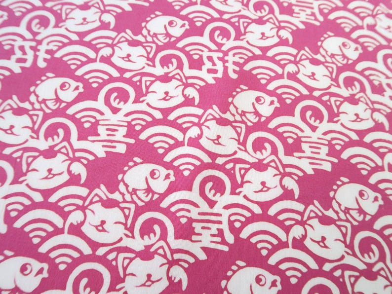 Pu&ograve; includere: Un tessuto rosa vivace con un motivo bianco ripetuto di gatti portafortuna stilizzati, pesci e onde. Il design include piccoli caratteri cinesi intervallati tra i gatti e i pesci, creando una stampa grafica densa e continua su tutto il tessuto.