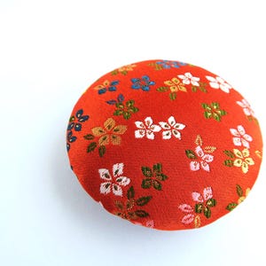 May include: A round, fabric-covered button featuring a vibrant orange background with a repeating pattern of small, multi-colored floral motifs in shades of white, pink, yellow, and dark blue, with metallic gold-toned thread accents throughout.