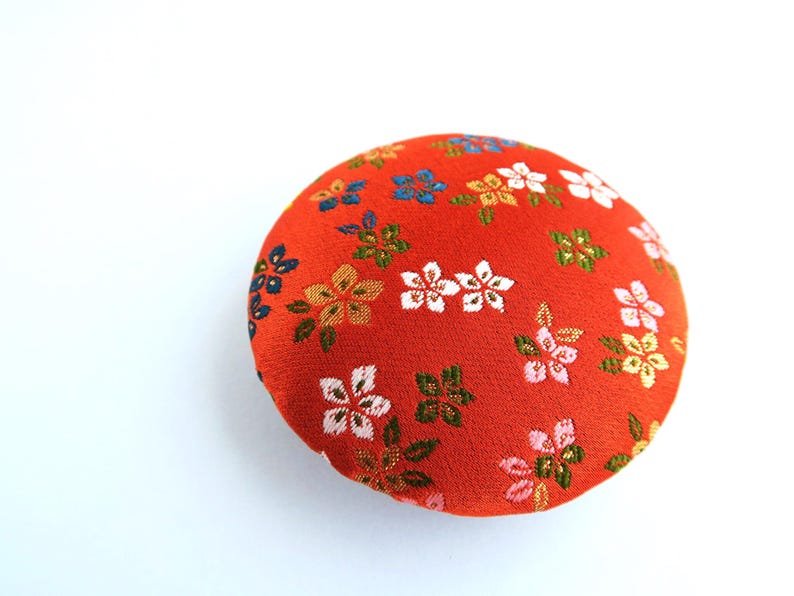May include: A round, fabric-covered button featuring a vibrant orange background with a repeating pattern of small, multi-colored floral motifs in shades of white, pink, yellow, and dark blue, with metallic gold-toned thread accents throughout.