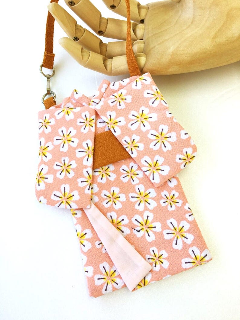 May include: A small pouch shaped like a kimono with a peach-colored base featuring a white and yellow floral pattern. The pouch includes a brown strap attached with a silver metal ring and a solid brown band across the center of the garment.