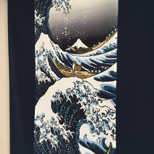 May include: A blue and white fabric wall hanging featuring a stylized wave with a dog standing on a boat. The wave is reminiscent of the Great Wave off Kanagawa by Hokusai. The background is a light gray with a mountain in the distance.