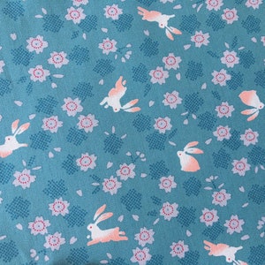 Peut inclure: Tissu bleu canard avec un motif r&eacute;p&eacute;t&eacute; de lapins blancs avec des accents p&ecirc;che et de petites fleurs roses et blanches. Les fleurs ont un centre rouge et sont dispers&eacute;es sur le tissu. Les lapins sont dans diverses poses.