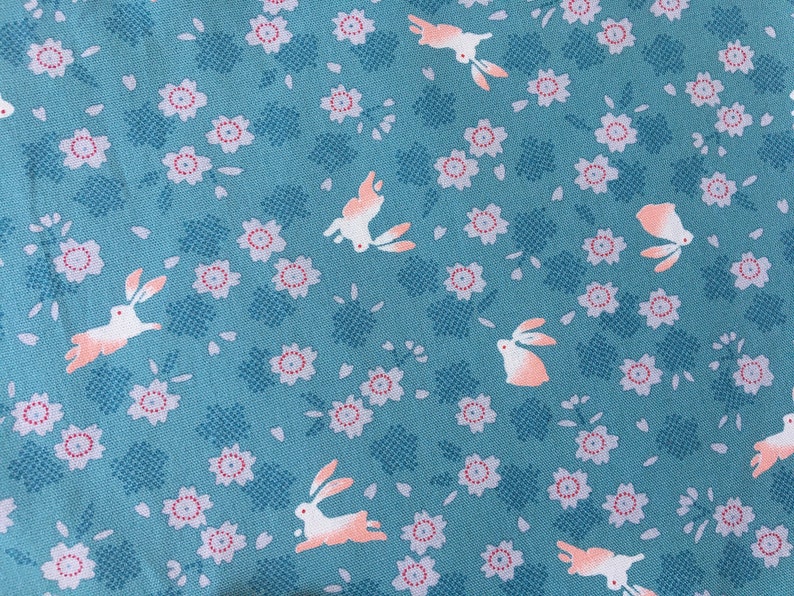 Peut inclure: Tissu bleu canard avec un motif r&eacute;p&eacute;t&eacute; de lapins blancs avec des accents p&ecirc;che et de petites fleurs roses et blanches. Les fleurs ont un centre rouge et sont dispers&eacute;es sur le tissu. Les lapins sont dans diverses poses.
