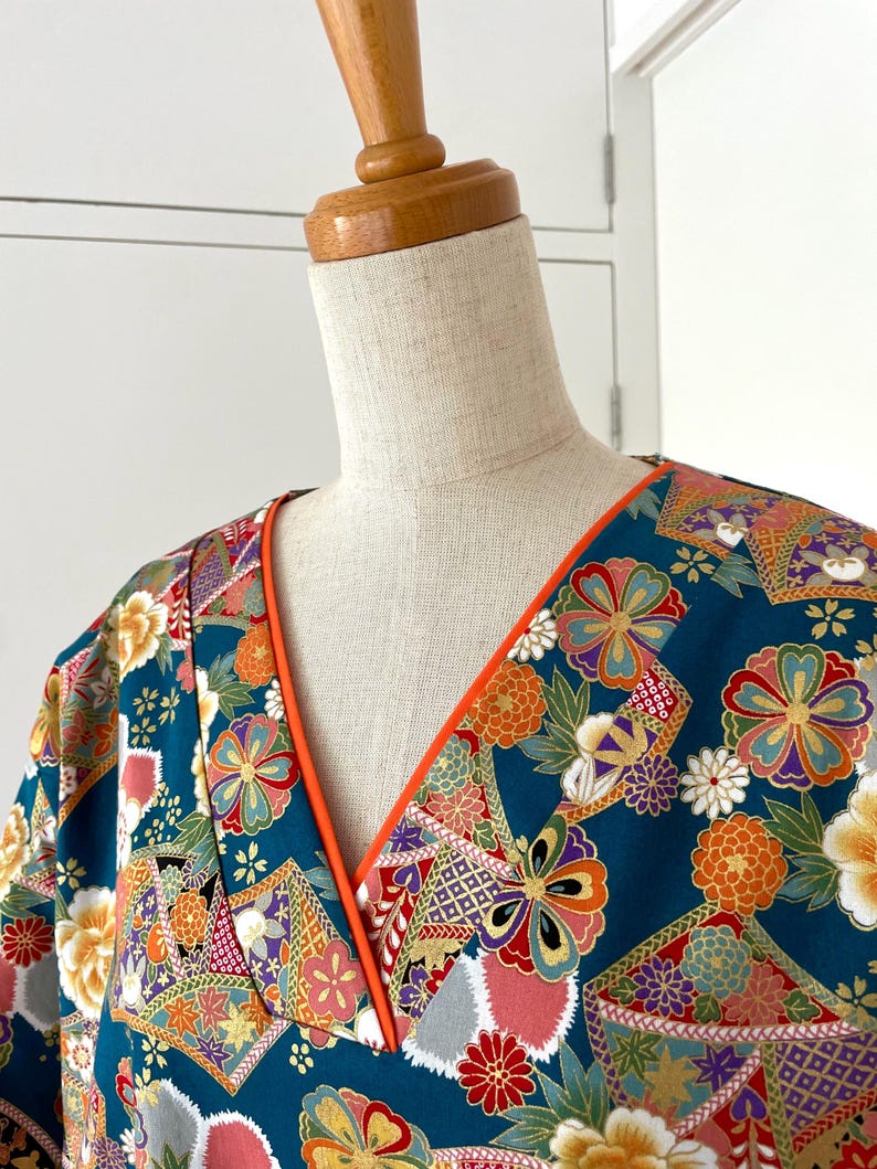 Puede incluir: Un kimono azul turquesa con un estampado floral colorido y ribete naranja alrededor del escote. El kimono est&aacute; en un maniqu&iacute; blanco.