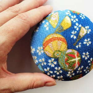 May include: A circular fabric-covered button featuring a vibrant blue background with a repeating pattern of small white cherry blossoms, yellow fans, and colorful spherical ornaments with red, green, and gold accents across the textured surface.