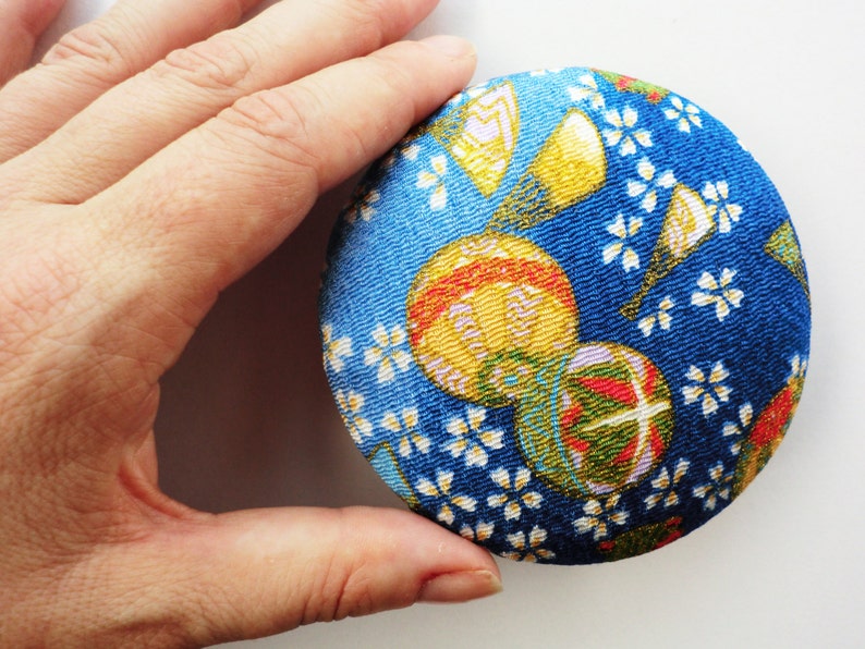May include: A circular fabric-covered button featuring a vibrant blue background with a repeating pattern of small white cherry blossoms, yellow fans, and colorful spherical ornaments with red, green, and gold accents across the textured surface.