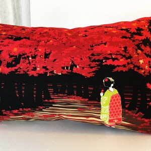 May include: A decorative pillow with a black background and a red and orange Japanese-inspired design. The design features a woman in a traditional kimono walking through a forest of red maple trees.