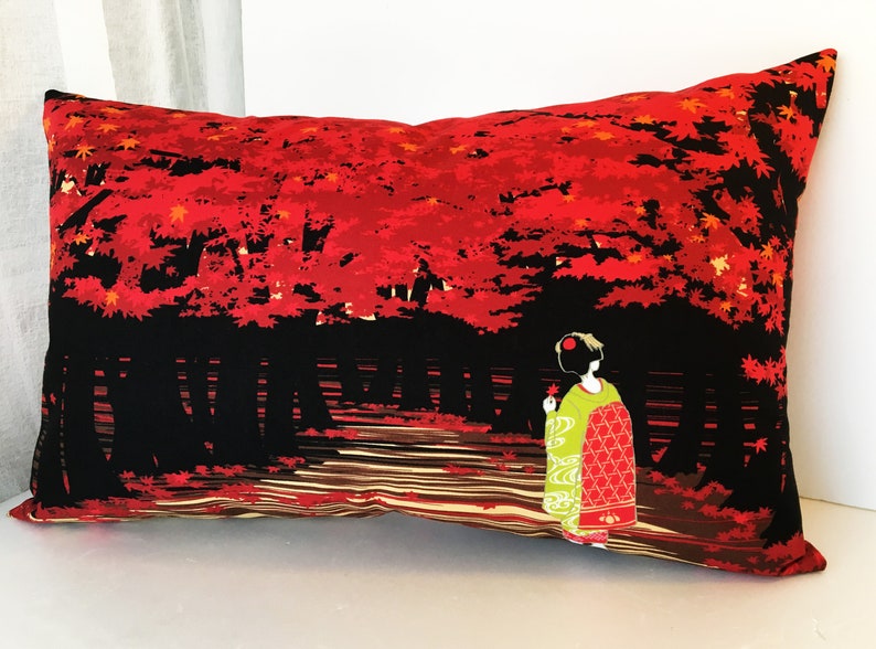 May include: A decorative pillow with a black background and a red and orange Japanese-inspired design. The design features a woman in a traditional kimono walking through a forest of red maple trees.