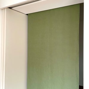 Puede incluir: Un divisor de habitaci&oacute;n de tela verde cuelga de un sistema de rieles. El divisor est&aacute; hecho de una tela texturizada y est&aacute; parcialmente abierto, revelando una pared blanca detr&aacute;s.